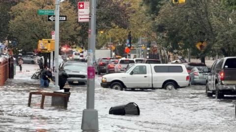 A long shot down a flooded street shows people in knee-high water and vehicles up to the tyres.