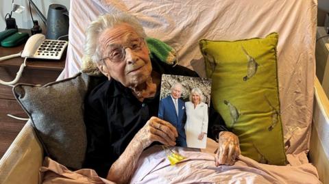 Marjorie Hodnett, a elderly lady, lies propped up in a bed, with two pillows either side of her. She has grey hair and is wearing glasses and holds up a picture of King Charles and Queen Camilla. 