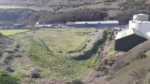 A large field where silt is deposited from the marina, there are industrial looking sheds at the back and on the right of the field.