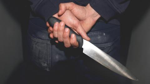 A man dressed in jeans and a dark top holding a knife behind his back. He is in a dark room.