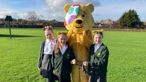 Three girls wearing yellow bear ears stand next to Pudsey in a field. Two of the girls are holding tights which tethered their legs together on the walk.