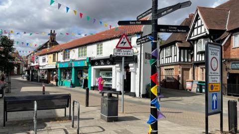 A view of the high street in Hessle. Rainbow bunting can be seen around the sign post directing to the town hall and other locations, as well as crisscrossing the street above the shops. 