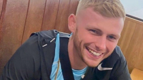Cody Fisher, a young man with blond hair, is wearing a light blue t-shirt with a dark tracksuit top over it. He is smiling at the camera.

