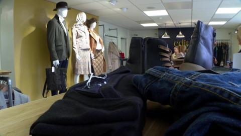 The inside of a new charity shop. A jumper, pair of jeans and boots are on a stand in the foreground. On the left are three mannequins, one male and two female, dressed in second-hand designer clothes. Further away a woman can be seen at the till in front of a sign with the shop's logo, the word 'Edit'.