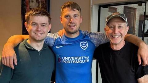 Three men standing in front of a glass door to a picture lined hallway. The man in the middle has short dark hair and a blue shirt, with his arms over the shoulders of the two other men. The man to the left also has short dark hair and is wearing a green shirt. The man to the right is wearing a cap and a black t-shirt.