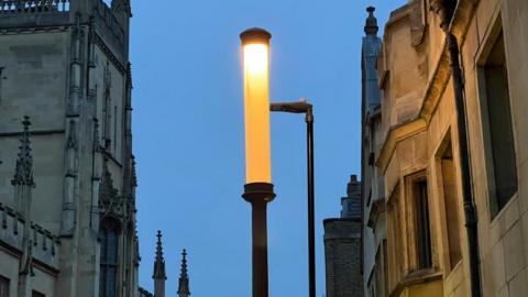 A lamp lit up against the early evening sky. It is tall and thin and there are ornate university buildings on either side