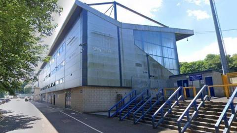 The sun shines above the main stand of the stadium, which is grey with blue pillars on top of light brown bricks.  In front of it are steps with thick blue bannisters leading up to the turnstiles, which have blue doors.