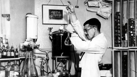 A man in a white coat with glasses and short black hair. He is stood in a room mixing chemicals. This is a black and white image.