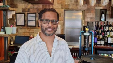 A man wearing a white shirt with black pinstripes stood in a pub.