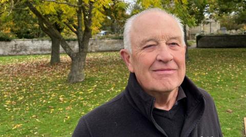 Gerry Kirt has grey hair on the very top of his head and down the sides of his head to his ears, he is wearing a black fleece over a black 'v' necked jumper, behind him is a village green, with trees and a village sign saying Glinton, a church wall is beyond that with gravestones and part of a church visible.