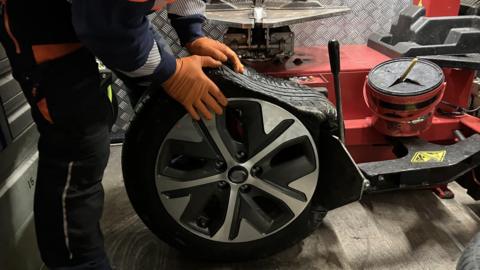 A close-up of a punctured tyre that is flat across the top. It is being held by a man wearing orange plastic gloves, inside his recovery van. His head and shoulders are not in shot.
