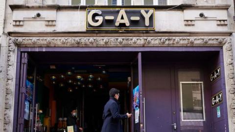 The entrance of G-A-Y Bar with purple walls and doors and the venue's name above it. A person can be seen walking past it in the daytime and a person is inside the club's entrance.