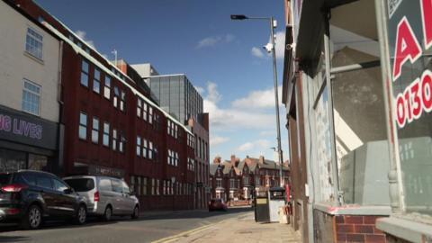 A view of a residential and retail street, with pavements and a road through the middle. Two cars are parked on the left hand side. 