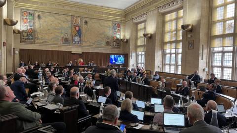 The Essex County Council chamber, with a meeting of councillors taking place. They are all sitting down on the benches, with one standing addressing the group with his back turned. There are large maps of Essex printed on the high walls.