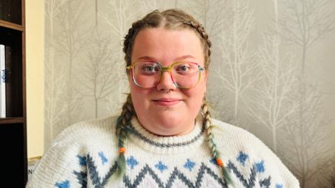 Samantha is sitting in a 'Welcome Space'. She is smiling a little and wearing a thick woolly jumper and glasses. There is wallpaper of winter trees behind her and a bookshelf beside her.    