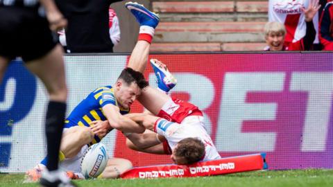 Hull KR player Joe Burgess, in white shirt and red shorts, flattens the corner post as he dives, legs in the air, after scoring a try as Warrington's Josh Thewlis, in blue and yellow hoops, tries to grab him