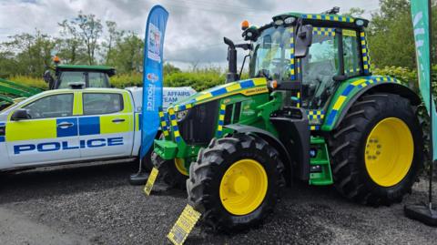 A green tractor with yellow and blue police stipes. In is parked next to a white blue and yellow police car. There are trees in the background. The sky is grey and cloudy.