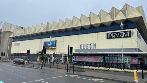 The south wall of the Kingwest building in Brighton. It is a white building with large white triangles on the roof. The word PRYZM and ODEON are written on the front. There is a horizontal blue stripe around the top quarter of the building.