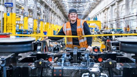 A woman is seen in a manufacturing facility, wearing an orange high visibility jacket and standing in front of a train chassis. She is in a brightly-lit warehouse with numerous work stations.