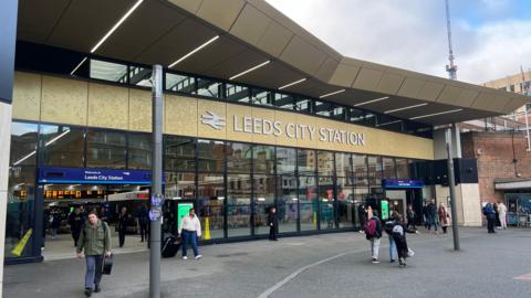 Outside Leeds City station. The building has a glass front, with the name of the station above the glass. It is a sunny day and people are walking in and out of the station. 