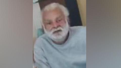 Sukhjinder Singh-Gill: A man with white hair and and white beard wearing a pale grey, round-necked, long-sleeved, top.

