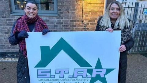 The two co-CEOs of STAR Housing smile at the camera whilst holding a large printed sign that features their company's green and pink logo on a white background. Chloe Hollingworth is on the left, has dark hair and is wearing a checked scarf. Zena Browne is on the right, has blond hair, a dark coat and a leopard skin patterned scarf.