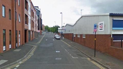 A street in Birmingham. It is quiet and there are only a couple of cars lining the road. On one side there are residential apartments. On the other there are industrial units.