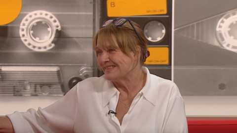 A woman in a white shirt sits on a red sofa with a background showing pictures of cassettes - the mix tapes - behind her. 