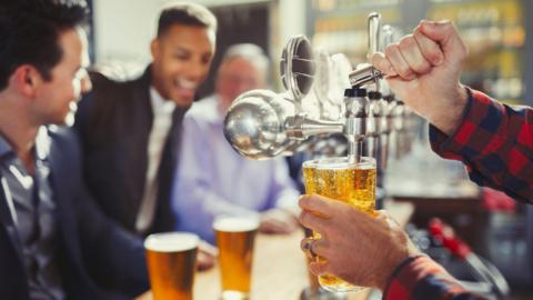 Close up of a pint being pulled from a tap. In the background there are blurred images of young men in shirts and jackets sitting at the bar with full pints in front of them. They look cheerful.