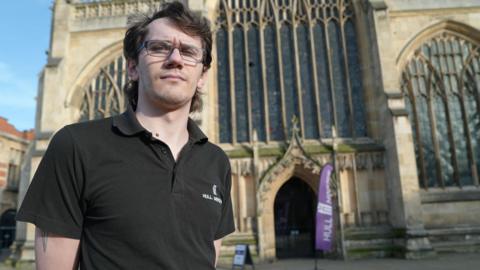 James Cameron - a man in a polo neck top with glasses and light brown hair. He looks frustrated and disheartened. He is standing in front of Hull Minster with a circular hole high up in the top left area of the stained-glass. 