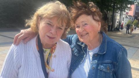 Two ladies stand in Derry city centre in an embrace. The lady on the right is wearing a white cardigan and is holding a black bag. she has shoulder length brown hair . To her right another lady is smiling, she has her arm around her companion. She is wearing a denim jacket and a white tshirt. Both ladies are standing in front of Derry's walls