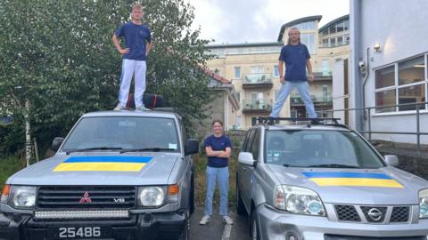 Fergus MacInnes and Emile Gonder standing on top of the two converted vehicles, with Daisy Livesey stood between the cars