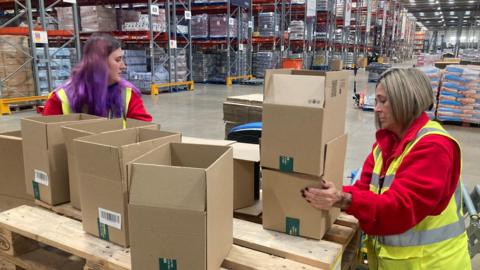 Two women in high visibility jackets pack cardboard boxes on top of a wooden pallet. They are in a vast warehouse environment. 