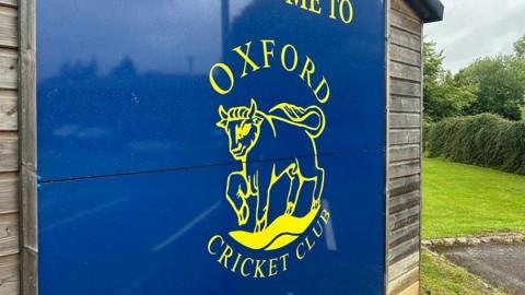 A blue sign with the Oxford Cricket Club logo, which is an ox, in yellow.