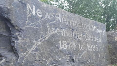 Grey memorial stone in front of trees with faded lettering that says New Herrington Miners’ Memorial Garden 1874 to 1985.