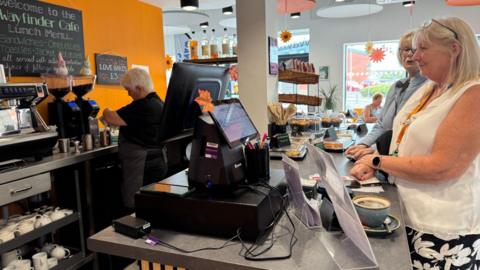 A photograph of the interior of a cafe. Two women stand behind the counter while a member of staff prepares a coffee. There are cakes and cups on the counter.