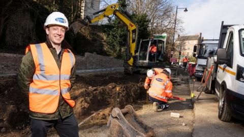 Councillor Tim McGuinness is wearing white hat, orange hi-vis vest, green jacket and dark blue jeans. He is stood in front of a newly developed bus stand near North Road in Durham City. There are two other workers with similar clothing, a yellow bulldozer digging and a white van and a white trucked parked on the kerb,