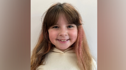 A young girl wearing a cream hoodie with brown long hair, with a faded pink streak to her left hand side of her hair, smiles at the camera