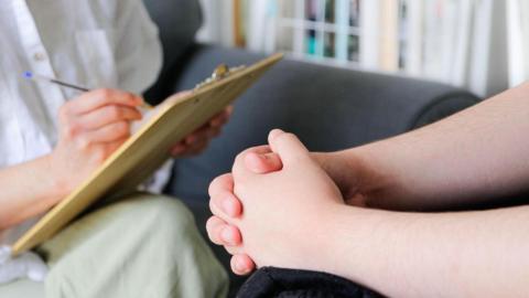 A generic image of a person having counselling with someone else holding a clipboard. We see two clasped hands as the person with the clipboard writes