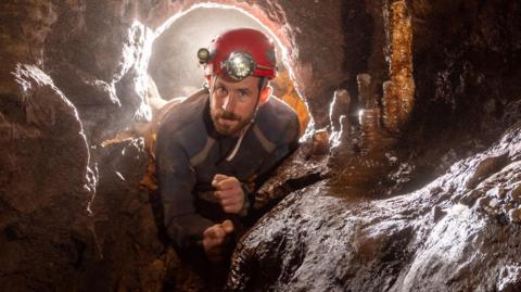 John Bolton, a young man with a brown beard and blue eyes is wearing a red helmet with a head torch. He is in a cave, with stalagmites to his side.