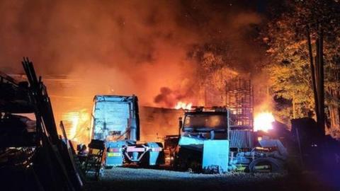 Lorries parked in front of building on fire with flames and smoke filling the air and trees surrounding