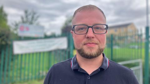 A man with blonde hair looks at the camera. He wears glasses and a dark blue polo shirt. Behind him, a Little Owls nursery setting is blurred.