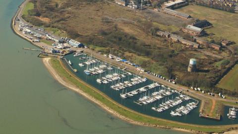 An aerial view of a marina full of boats. The lock to the marina is on the left with buildings, a car park next to it. A road is behind the marina, with scrubland beyond it, which has a water tower on it and former naval base buildings.