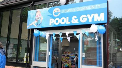 Elswick Community Pool and Gym. The glass fronted building has a large blue sign hanging above its door. Balloons and bunting are displayed above the entrance to celebrate its reopening.