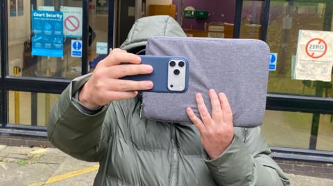 A man wearing a dark green coat with a laptop in his left hand covering his face and his right hand holding a phone to the camera. He is stood in front of black sliding doors, the main entrance to Guildford Magistrates' Court.