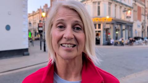 A woman with short, blonde hair standing in the street, smiling at the camera. She is wearing a red jacket. 