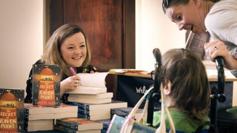 Author Lisette Auton at a book signing. She sits behind a desk covered in books while a child and parent wait for a signed copy of one of her books.