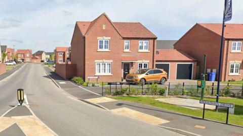 Street view of Berriman Drive from a roundabout. On the left is the rear of a 'keep left' bollard on a pedestrian island, which is in the middle of the entrance to the road. In the far distance is a row of new-build houses. On the right is a red-brick double-fronted house with a gold-coloured SUV parked in the driveway. In front of the property is a very small public green space with a blue flag erected in the middle.