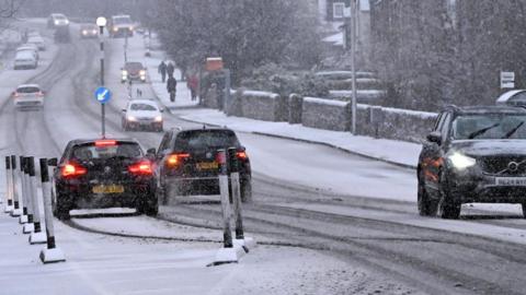 Cars driving on a road with snow falling and starting to settle on the pavement and partially the road itself
