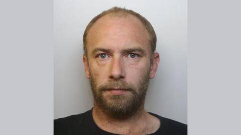 A police custody photo of Adam Dodden. He has short brown hair and a short beard. He wears a black t-shirt.